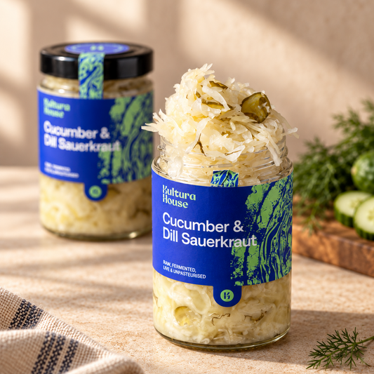 Two glass jars of Natura House Cucumber & Dill Sauerkraut on a beige fabric surface with a striped cloth, fresh dill, and sliced cucumbers in the background.